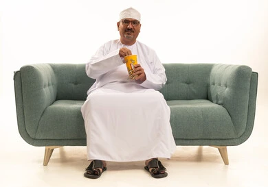الاستمتاع بتناول الفشار، انشطة ترفيهية منزلية، بورتريه كبير مسن لرجل عربي خليجي عماني يرتدي الدشداشة والكمة التقلييدية يحمل بيده علبة الذرة، سناكات ومقرمشات لذيذة، تناول سناكات خفيفة وصحية، الاستمتاع باكل البوب كورن، الترفيه عن النفس وقضاء وقت ممتع،خلفيه بيضاء