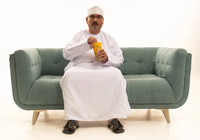 انشطة ترفيهية منزلية، الاستمتاع بتناول الفشار، بورتريه كبير مسن لرجل عربي خليجي عماني يرتدي الدشداشة والكمة التقلييدية يحمل بيده علبة الذرة، سناكات ومقرمشات لذيذة، تناول سناكات خفيفة وصحية، الاستمتاع باكل البوب كورن، الترفيه عن النفس وقضاء وقت ممتع،خلفيه بيضاء