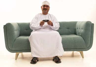 انشطة ترفيهية منزلية، ايماءات الفوز والحماس، قضاء وقت ممتع في المنزل، استخدام التكنولوجيا الحديثة في الترفيه، بورتريه  لرجل مسن عربي خليجي عماني يرتدي الدشداشة والكمة يجلس على الأريكه يمسك بيده عصا التحكم ، المتعة والتسلية والتفاعل، خلفية بيضاء