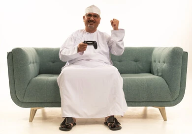 حركة يد ايماءات فوز وحماس، قضاء وقت ممتع في المنزل، انشطة ترفيهية منزلية، استخدام التكنولوجيا الحديثة في الترفيه، بورتريه  لرجل مسن عربي خليجي عماني يرتدي الدشداشة والكمة يجلس على الأريكه يمسك بيده عصا التحكم ، المتعة والتسلية والتفاعل، خلفية بيضاء