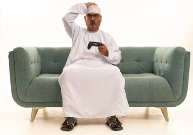 إيماءات الحزن والخسارة، قضاء وقت ممتع في المنزل، انشطة ترفيهية منزلية، استخدام التكنولوجيا الحديثة في الترفيه، بورتريه لرجل مسن عربي خليجي عماني يرتدي الدشداشة والكمة يجلس على الأريكه يمسك بيده عصا التحكم ، المتعة والتسلية والتفاعل، خلفية بيضاء