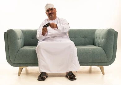 قضاء وقت ممتع في المنزل، انشطة ترفيهية منزلية، استخدام التكنولوجيا الحديثة في الترفيه، بورتريه لرجل كبير مسن عربي خليجي عماني يرتدي الدشداشة والكمة يجلس على الأريكه يمسك بيده عصا التحكم يقوم بلعب العاب الفيديو بإيماءات السعادة والسرور، المتعة والتسلية والتفاعل، خلفية بيضاء