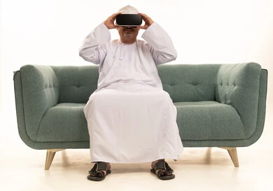 Integrating modern technologies with daily life, gestures of interaction and integration, spending entertaining times with virtual games, a portrait of an elderly Arab Gulf Omani man wearing a traditional dishdasha and kumma sitting on a comfortable sofa and wearing 3D virtual reality glasses, white background.