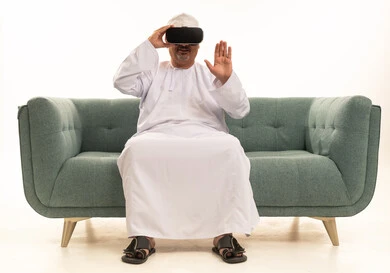 التفاعل والاندماج مع العناصر الهولوجرامية، قضاء أوقات ممتعة ومسلية بالألعاب الافتراضية، بورتريه لرجل عربي خليجي عماني كبير بالسن يرتدي الدشداشة والكمة التقليدية يجلس على آريكة مريحة  ويرتدي نظارة الواقع الافتراضي ثلاثية الابعاد،  خلفية بيضاء