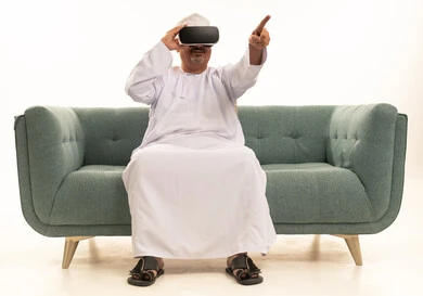 Gestures of interaction and integration with holographic elements, spending enjoyable and entertaining times with virtual games, a portrait of an elderly Arab Gulf Omani man wearing a traditional dishdasha and kumma sitting on a comfortable sofa and wearing 3D virtual reality glasses, white background.