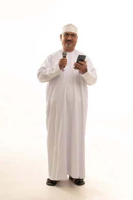 تقديم فقرات ثقافية، مهارة وفن الألقاء، مفهوم الرجولة والوقار، بورتريه لرجل كبير بالسن عربي خليجي عماني يرتدي الدشداشة التقليدية والكمه العمانية والنظارة الطبية يقف ويحمل مايكروفون وهاتف بإيماءات البشاشة والسرر، الجسم بالطول الكامل، خلفية بيضاء