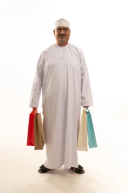 Gestures of happiness and joy at discounts, benefiting from seasonal discounts, buying products and items, a portrait of an elderly Arab Gulf Omani man wearing a traditional dishdasha and kumma holding paper bags and a mobile phone, a full-body image, white background.