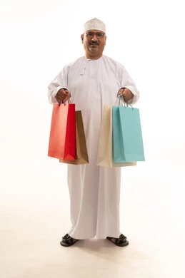 Buying products and items, gestures of happiness and joy at discounts, taking advantage of seasonal discounts, a portrait of an elderly Arab Gulf Omani man wearing a traditional dishdasha and kumma holding paper bags and a mobile phone, full-body image, white background.
