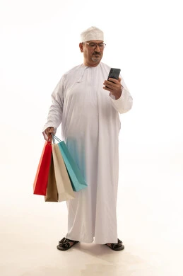 The use of technology in daily life, buying products and items online, a portrait of an elderly Arab Gulf Omani man wearing a traditional dishdasha and kumma holding paper bags and a mobile phone, a full-body image, white background.