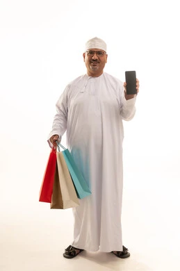 Displaying a blank black screen on the mobile, buying products and items online, a portrait of an elderly Arab Gulf Omani man wearing a traditional dishdasha and kumma holding paper bags and a mobile phone, a full-body image, white background.