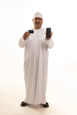 Using mobile applications for payment and shopping, a portrait of an elderly Arab Gulf Omani man wearing a dishdasha and a kumma, holding an electronic card and a mobile phone, looking with gestures of cheerfulness and joy, the possibility of electronic payment through advanced modern technologies, white background.