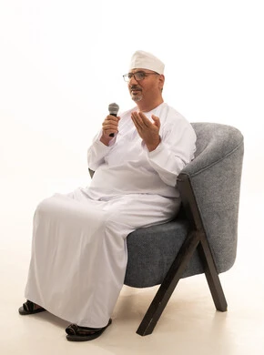 القاء الخطابات والكلمات باستخدام مكبر الصوت ، تسجيل المقاطع الصوتية والغنائية، تبادل الحوار والاستفادة من خبرات كبار السن، بورتريه لرجل عربي خليجي عماني يرتدي الدشداشة والعمامة يجلس على كرسي مريح يحمل بيده مكبر الصوت، صورة الجسم بالطول الكامل، خلفية بيضاء