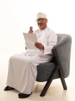 Exchanging dialogue and benefiting from the experiences of the elderly, conducting interviews and asking questions, engaging in discussions and debates, delivering speeches and words, a portrait of an Arab Gulf Omani man wearing a dishdasha and a turban sitting on a comfortable chair holding a microphone and a white paper, full-length image, white background.