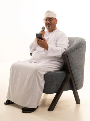 Entertainment segments and presenting theatrical performances, interviews and asking questions, conducting dialogues and discussions, delivering speeches and words, a portrait of an Arab Gulf Omani man wearing a dishdasha and turban sitting on a comfortable chair holding a megaphone in his hand, full-length image, white background.