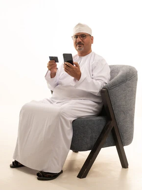 إمكانية تسديد الفواتير بالبطاقات الإلكترونية, استخدام جهاز تقني حديث, سهولة الشراء والدفع عن طريق بطاقة الإئتمان البنكية، بورتريه لرجل مسن عربي خليجي عماني يرتدي دشداشة وكمة ونظارة طبية يجلس على كرسي ويمسك بيده بطاقة الصراف ويعمل على الهاتف المحمول، خلفية بيضاء