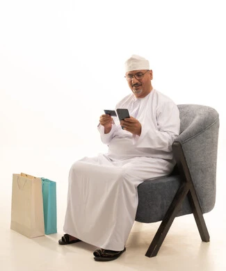 إمكانية التسوق والتبضع عبر الانترنت, استخدام جهاز تقني حديث, سهولة الشراء والدفع عن طريق بطاقة الإئتمان البنكية، بورتريه لرجل مسن عربي خليجي عماني يرتدي دشداشة وكمة ونظارة طبية يجلس على كرسي ويمسك بيده بطاقة الصراف ويعمل على الهاتف المحمول، خلفية بيضاء