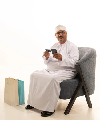 The possibility of shopping and purchasing online, using modern technology, ease of buying and paying with a bank credit card, a portrait of an elderly Arab Gulf Omani man wearing a dishdasha and a kumma and medical glasses sitting on a chair holding an ATM card and working on a mobile phone, white background.