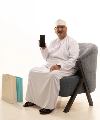 A display of a blank black screen on a mobile phone, the ability to shop online, paper shopping bags, the use of modern technology, a portrait of an elderly Arab Gulf Omani man wearing a dishdasha and kumma and medical glasses sitting on a chair and working on a mobile phone, white background.