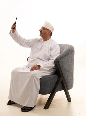 التقاط صورة سيلفي تذكارية عبر الجوال، استخدام جهاز تقني حديث، بورتريه لرجل كبير السن عربي خليجي عماني يرتدي دشداشة وكمة ونظارة طبية يجلس على كرسي مريح وحمل بيده الهاتف المحمول، خلفية بيضاء