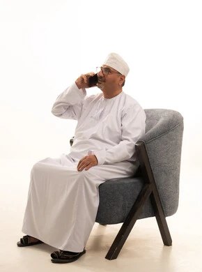 Communicating with family and friends, making a phone call via mobile, using modern technology, a portrait of an elderly Arab Gulf Omani man wearing a dishdasha and kumma and reading glasses sitting on a comfortable chair and working on a mobile phone, white background.