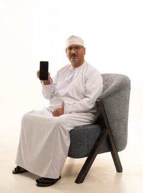 عرض شاشة سوداء فارغة عبر الجوال، استخدام جهاز تقني حديث، بورتريه لرجل مسن عربي خليجي عماني يرتدي دشداشة وكمة ونظارة طبية يجلس على كرسي ويمسك بيده بطاقة الصراف ويعمل على الهاتف المحمول، خلفية بيضاء
