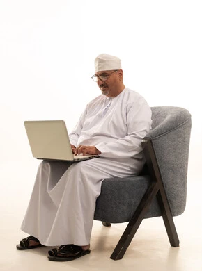 Completing work tasks via laptop, using a modern technical device, a portrait of an elderly Arab Gulf Omani man wearing a dishdasha and kumma and reading glasses, sitting on a comfortable chair and working on a laptop, white background.