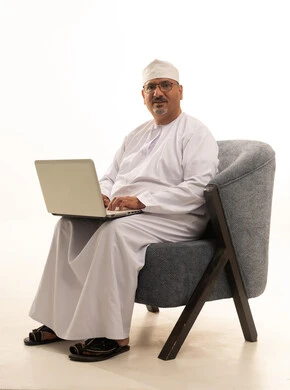 Completing work tasks via laptop, using a modern technical device, a portrait of an elderly Arab Gulf Omani man wearing a dishdasha and kumma and reading glasses, sitting on a comfortable chair and working on a laptop, white background.