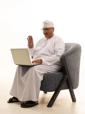 القاء التحية والسلام، اجراء مكالمة فيديو، إنجاز مهام العمل عبر اللابتوب، استخدام جهاز تقني حديث، بورتريه لرجل كبير السن عربي خليجي عماني يرتدي دشداشة وكمة ونظارة طبية يجلس على كرسي مريح ويعمل على الحاسوب المحمول، خلفية بيضاء