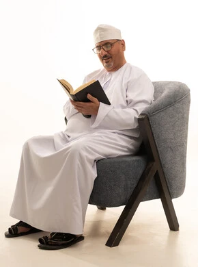 Gathering information from books and novels, the concept of self-learning, expressions of contemplation and focus, spending time reading, learning, and culture, a portrait of an elderly Arab Omani Gulf man wearing a dishdasha and kumma and reading glasses sitting on a comfortable chair reading a book, white background.