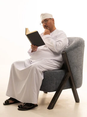 Gathering information from books and novels, the concept of self-learning, expressions of contemplation and focus, spending time reading, learning, and culture, a portrait of an elderly Arab Omani Gulf man wearing a dishdasha and kumma and reading glasses sitting on a comfortable chair reading a book, white background.