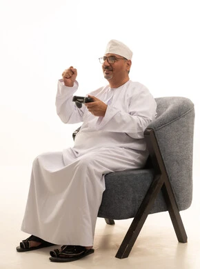 Spending enjoyable and entertaining time, fun modern technologies, interaction and immersion with PlayStation games, a portrait of an elderly Arab Omani Gulf man wearing a dishdasha and kumma and medical glasses sitting on a comfortable chair holding a game controller and playing video games, white background.