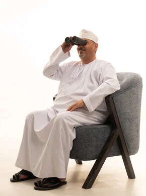 Future visions among adults and the elderly, a love for exploration and adventure, a portrait of an elderly Arab Gulf Omani man smiling, wearing a dishdasha, a kumma, and reading glasses, looking through a binocular telescope, seeing distant objects, with a white background.