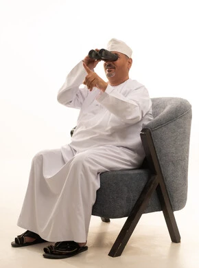 حب الاستكشاف والمغامرة، الإشارة بإصبع السبابة إلى شيء ما، رؤى مستقبلية عند الراشدين والكبار بالسن، بورتريه لرجل كبير مسن عربي خليجي عماني مبتسم يرتدي دشداشة وكمة ونظارات طبية يقوم بالنظر من خلال المنظار ثنائي العينين(التلسكوب)، رؤية الأشياء البعيدة، خلفية بيضاء.