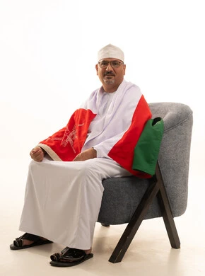 Looking at the camera with gestures of joy and happiness, cheering for the national team, a portrait of an elderly Arab Gulf Saudi man wearing a thobe and a headscarf, with glasses, sitting on a comfortable chair and draping the flag of Oman over his shoulders, watching sports matches, white background. Looking at the camera with gestures of joy and happiness, cheering for the national team, a portrait of an elderly Arab Gulf Saudi man wearing a thobe and a headscarf, with glasses, sitting on a comfortable chair and draping the flag of Oman over his shoulders, watching sports matches, white background.