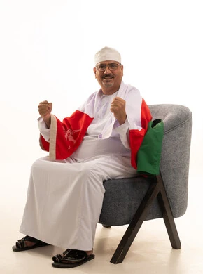 رفع قبضتي اليدين بإيماءات السرور والتشجيع، الحماس والتفاعل مع المنتخب الوطني، بورتريه لرجل كبير السن عربي خليجي سعودي يرتدي دشداشة وكمة ونظارات طبية يجلس على كرسي مريح ويضع علم سلطنة عمان على كتفيه، مشاهدة المباريات الرياضية، خلفية بيضاء