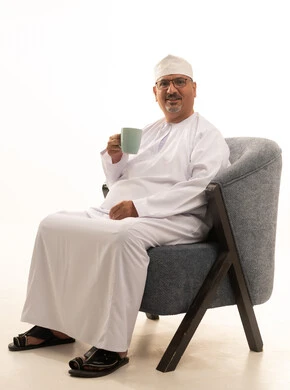 Sipping the favorite drink, enjoying warm winter herbs, relishing hot beverages, a portrait of a smiling Arab Gulf Omani man wearing a dishdasha, a kumma, and glasses sitting on a comfortable chair drinking coffee with gestures of happiness and joy, white background.