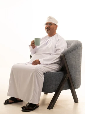 تناول الأعشاب الشتوية الدافئة، احتساء المشروب المفضل, الاستمتاع بالمشروبات الساخنة، بورتريه لرجل عربي خليجي عماني مبتسم يرتدي الدشداشة والكمة والنظارة الطبية يجلس على كرسي مريح يقوم بشرب القهوة بإيماءات السعادة والسرور، خلفية بيضاء