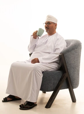 Sipping the favorite drink, enjoying warm winter herbs, relishing hot beverages, a portrait of a smiling Arab Gulf Omani man wearing a dishdasha, a kumma, and glasses sitting on a comfortable chair drinking coffee with gestures of happiness and joy, white background.
