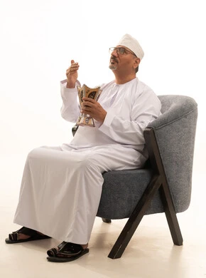 التعطر والتطيب بالبخور, استخدام خشب العود بالأعياد والمناسبات, العادات والتقاليد السعودية, بورتريه لرجل كبير مسن عربي خليجي عماني يرتدي الدشداشة والكمة والنظارات الطبية يجلس على كرسي مريح يقوم بالتعطر من المبخرة, الاهتمام بالمظهر الخارجي والأناقة, خلفية بيضاء