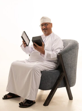 إيماءات السعادة والدهشة بالحصول على هدية، مفهوم المفاجأة والإهداء، فن انتقاء الهدايا بعناية، بورتريه لرجل مسن عربي خليجي عماني مبتسم  يرتدي الدشداشة والكمه والنظارات الطبية يجلس على كرسي مريح ويقوم بفتح العلبة بتعابير الفرح والسرور، خلفية بيضاء
