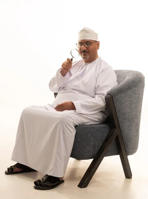 The concept of examination and exploration, clarifying the image and vision up close, a portrait of an elderly Arab Gulf Omani man wearing a dishdasha and kumma and medical glasses sitting on a comfortable chair holding a magnifying glass that he places on his eye with gestures of focus and contemplation, white background.