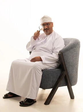 Clarification of the image and close-up vision, the concept of examination and exploration, a portrait of an elderly Arab Gulf Omani man wearing a dishdasha and kumma and medical glasses sitting on a comfortable chair holding a magnifying glass that he places on his eye with gestures of focus and contemplation, white background.