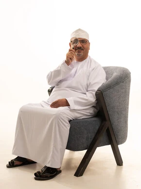 مفهوم الفحص والاستكشاف، توضيح الصورة والرؤية عن قرب، بورتريه لرجل كبير السن عربي خليجي عماني يرتدي الدشداشة والكمة ونظارات طبية يجلس على كرسي مريح يحمل بيده عدسة مكبرة يضعها على عينه بإيماءات التركيز والتمعن، خلفية بيضاء