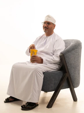 قضاء وقت الفراغ بمتابعة الافلام و المسلسلات المسلية، تناول سناك صحي و خفيف ، بورتريه لرجل كبير السن عربي خليجي عماني يرتدي الدشداشة والكمة ونظارات طبية يجلس على اريكه مريحة ويقوم بتناول الفشار، خلفية بيضاء