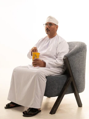 قضاء وقت الفراغ بمتابعة الافلام و المسلسلات المسلية، تناول سناك صحي و خفيف ، بورتريه لرجل كبير السن عربي خليجي عماني يرتدي الدشداشة والكمة ونظارات طبية يجلس على اريكه مريحة ويقوم بتناول الفشار، خلفية بيضاء