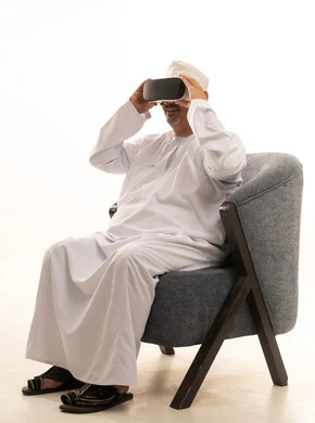 مفهوم التقنيات الإلكترونية الحديثة، التفاعل والاندماج مع العناصر الهولوجرامية، بورتريه لرجل كبير بالسن عربي سعودي خليجي يجلس على كرسي مريح يرتدي الدشداشة والكمة ونظارات الواقع الإفتراضي (في آر) ثلاثية الأبعاد، خلفية بيضاء
