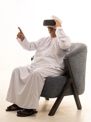 Interaction and integration with holographic elements, the concept of modern electronic technologies, a portrait of an elderly Arab Saudi Gulf man sitting on a comfortable chair wearing a thobe and a headscarf, and 3D virtual reality (VR) glasses, with a white background.
