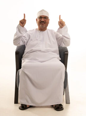 الاشارة بإصبع السبابة إلى شيء ما، النظر إلى الكاميرا بتعابير السعادة والسرور، بورتريه لرجل كبير مسن عربي عماني خليجي مبتسم يرتدي الدشداشة والكمه ونظارات طبية يجلس على كرسي مريح، خلفية بيضاء.