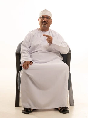 Pointing with the index finger at something, looking at the camera with expressions of happiness and joy, a portrait of an elderly Arab Omani Gulf man smiling, wearing a dishdasha and kumma, and wearing glasses, sitting on a comfortable chair, with a white background. Pointing with the index finger at something, looking at the camera with expressions of happiness and joy, a portrait of an elderly Arab Omani Gulf man smiling, wearing a dishdasha and kumma, and wearing glasses, sitting on a comfortable chair, with a white background.
