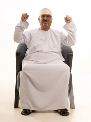 Raising his hands up in gestures of victory and triumph, looking at the camera with expressions of excitement and joy, a portrait of an elderly Arab Omani Gulf man smiling, wearing a dishdasha and kumma, and glasses, sitting on a comfortable chair, with a white background. Raising his hands up in gestures of victory and triumph, looking at the camera with expressions of excitement and joy, a portrait of an elderly Arab Omani Gulf man smiling, wearing a dishdasha and kumma, and glasses, sitting on a comfortable chair, with a white background.